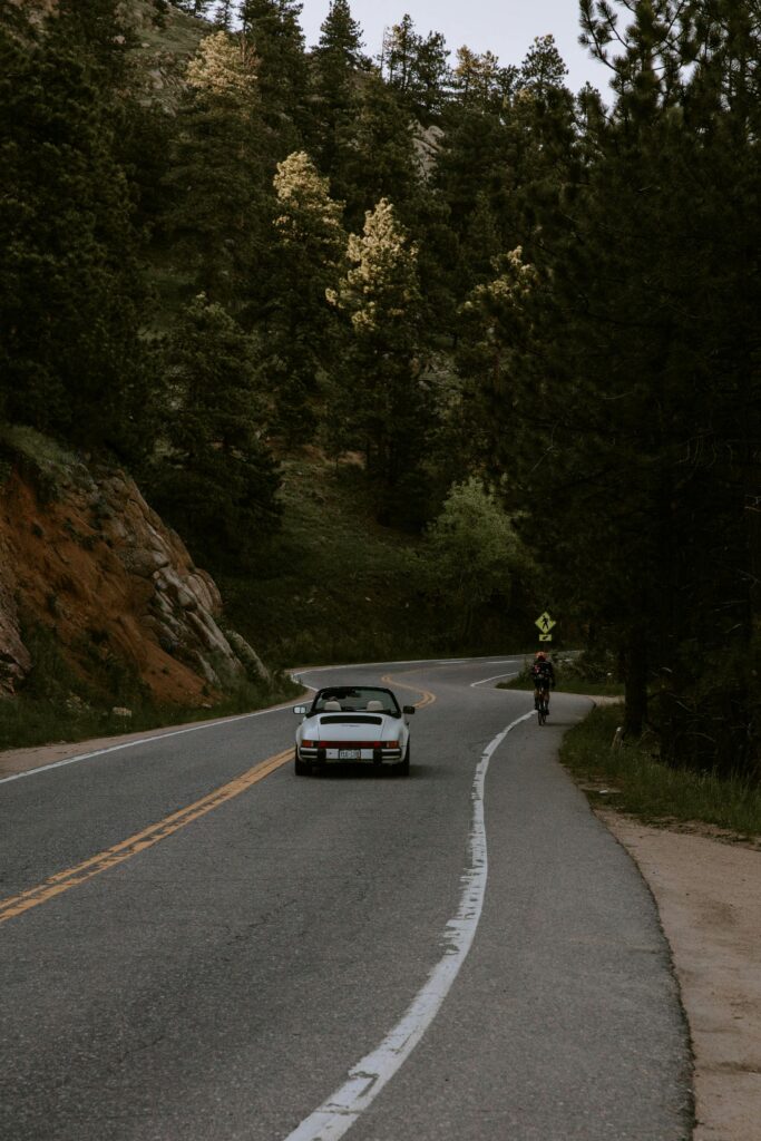 A car and cyclist on a winding mountain road lined with lush trees.